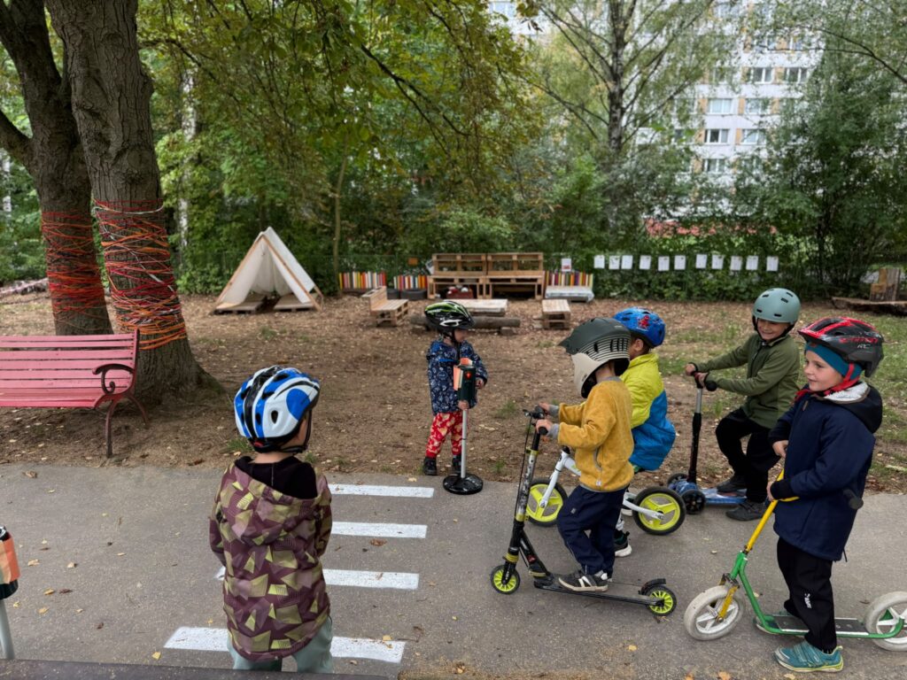 Děti s helmami na koloběžkách a odrážedlech čekají na hřišti mateřské školy před přechodem.