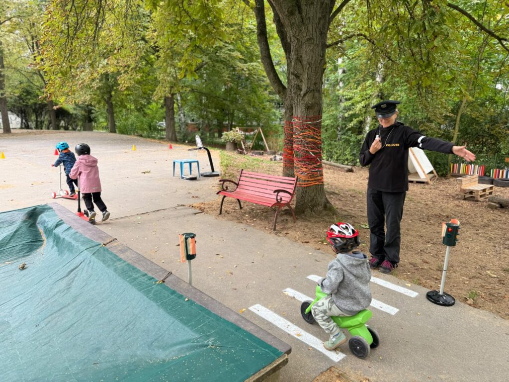 Paní policistka ukazuje dětem na hřišti, jak jezdit přes přechod; chlapec na zeleném odrážedle projíždí kolem semaforu.