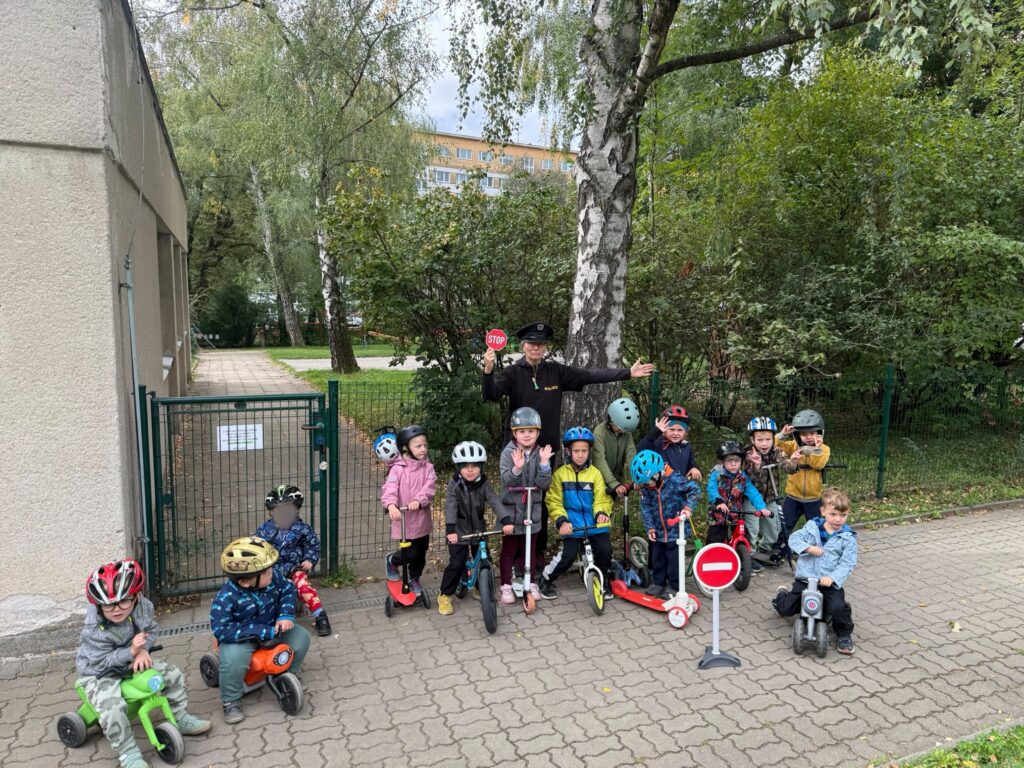 Děti s helmami na odrážedlech a koloběžkách se společně s paní policistkou fotí u branky mateřské školy.
