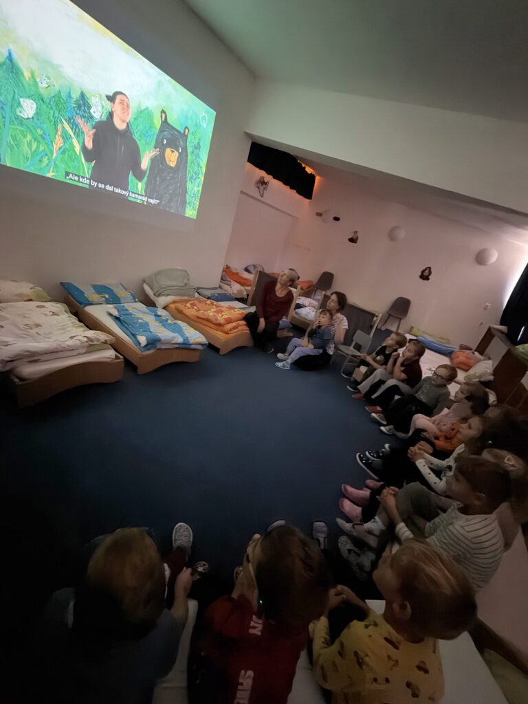 Děti sedí ve ztemnělé místnosti a sledují na stěně promítanou pohádku Kamarád pro medvěda.