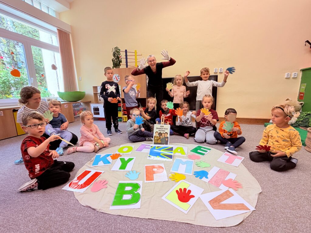 Děti sedí kolem velkého kruhu s barevnými písmeny abecedy a papírovými dlaněmi, paní učitelka a děti zvedají ruce.