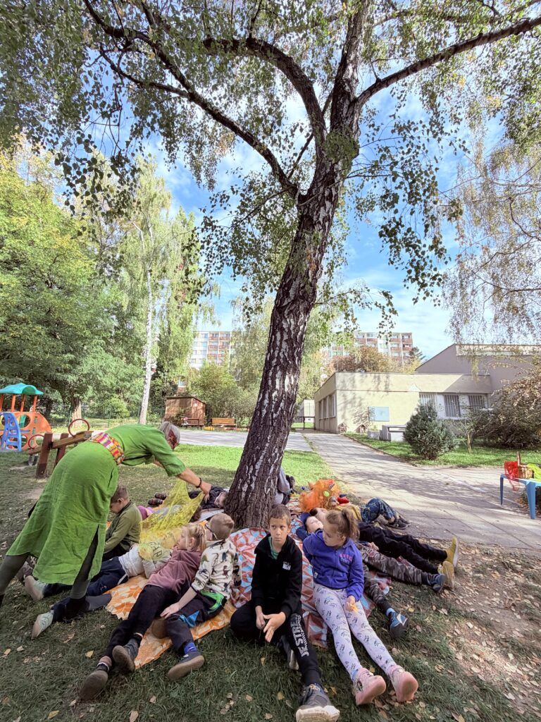Děti odpočívají na dekách pod velkou břízou a relaxují při hudbě.