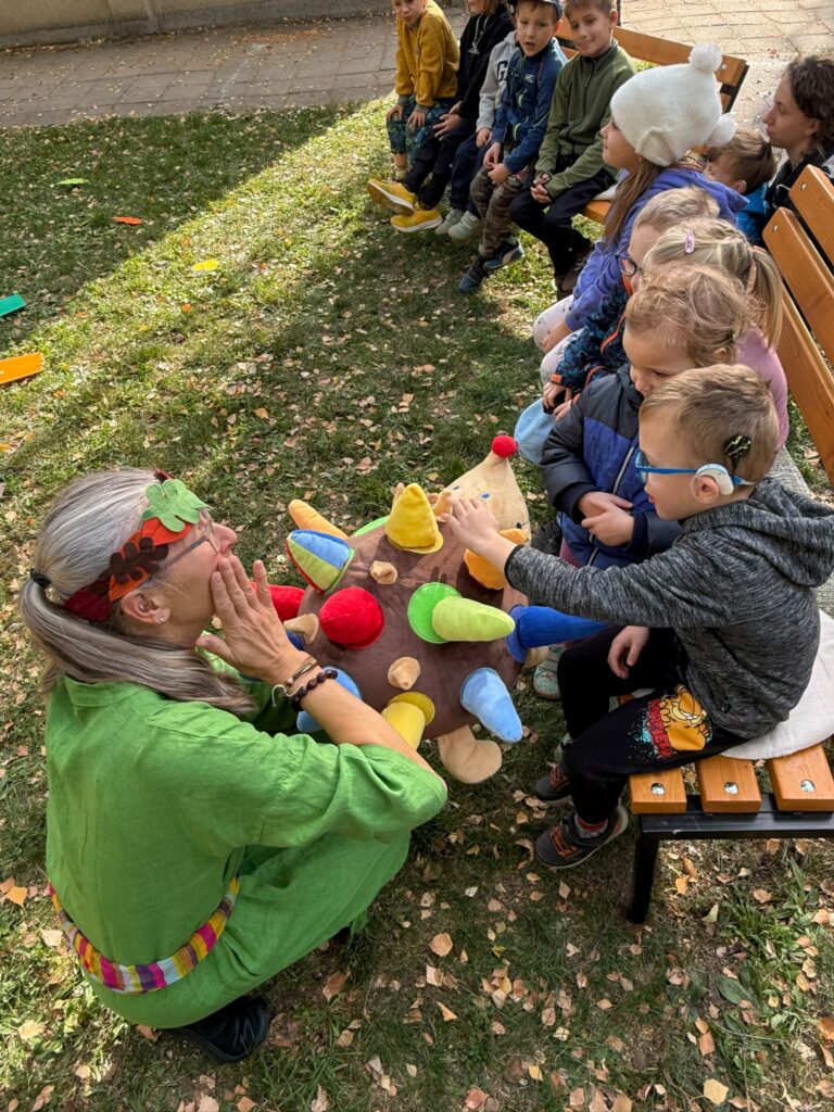 Herečka v zeleném kostýmu ukazuje dětem na lavičce velkého látkového ježka s barevnými ostny.