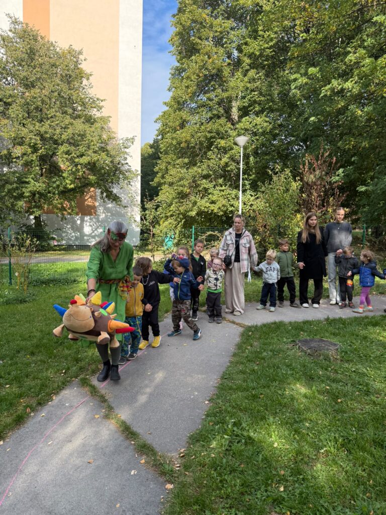 Děti se drží za ruce a putují s herečkou a látkovým ježkem po chodníčku zahradou.
