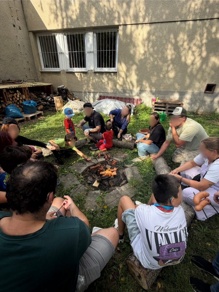 Děti, rodiče a pedagogové sedí na školní zahradě v kruhu kolem ohně a opékají špekáčky.