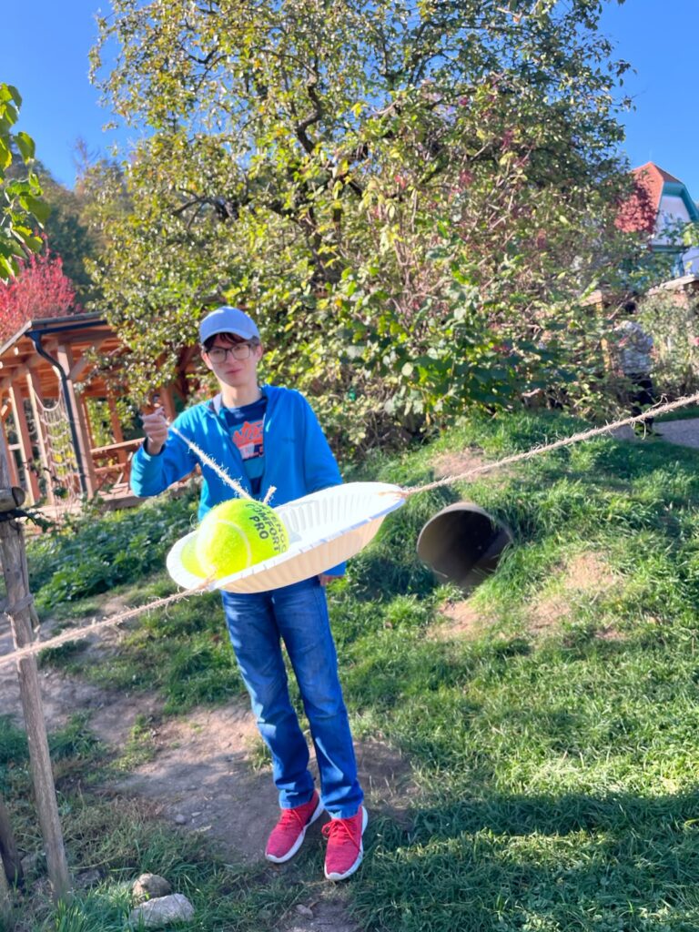 Žák v modré mikině navléká papírový talíř s tenisovým míčkem po napnutém provázku na zahradě.