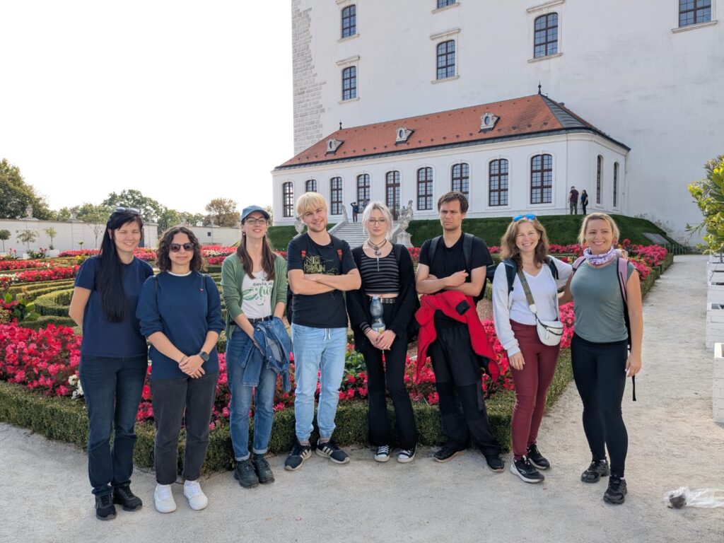 Skupinová fotografie maturantů s pedagožkou před Bratislavským hradem a zahradou s červenými květy.