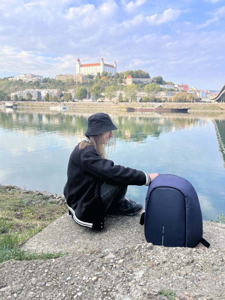 Studentka sedí na břehu Dunaje, v pozadí se nad řekou tyčí Bratislavský hrad.